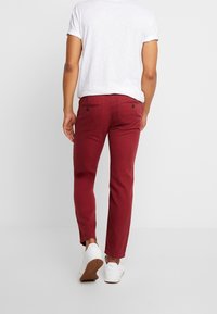 Man in een slim-fit bordeauxrode broek, een witte t-shirt met opgerolde mouwen en witte sneakers, die wegloopt tegen een effen achtergrond.