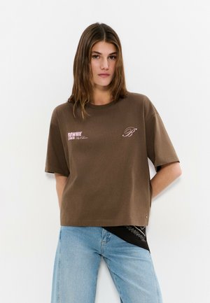 Mujer joven con cabello castaño, vestida con una camiseta marrón con diseño gráfico y vaqueros azul claro, posando frente a un fondo blanco liso.