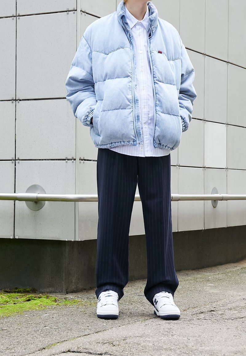 Personne portant une doudoune bleu clair, une chemise blanche boutonnée, un pantalon à fines rayures foncé et des baskets blanches, debout à l'extérieur près d'un mur carrelé.