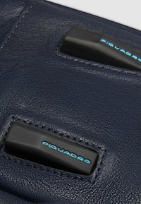 Superficie in pelle blu navy con due decorazioni nere con il logo "Piquadro" in blu. Texture liscia con dettagli di cuciture visibili.