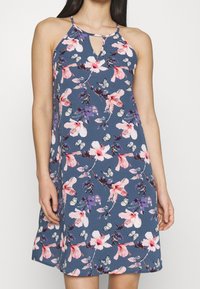 Blumendruckkleid mit Halterneck und Schlüssellochdetails. Aus weichem Stoff in Marineblau, mit rosa und lila Blumen und Schmetterlingen.