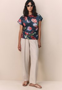 Haut bleu marine à motif floral en satin, associé à un pantalon large beige clair. Le modèle porte des sandales plates argentées et des lunettes de soleil rondes.