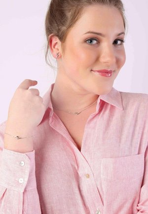 Collier et bracelet en argent avec des détails courbés, portés sur une chemise à rayures rose et blanche. Cheveux tirés en arrière, maquillage léger.