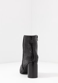 Bota negra hasta el tobillo con un diseño de tejido texturizado, cierre con cremallera y un tacón cilíndrico grueso. Material suave con un acabado brillante.
