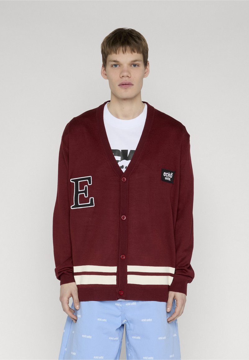 Νεαρός άνδρας φοράει ένα μπορντό ζακέτα με λευκές ρίγες και patches πάνω από ένα λευκό T-shirt και ανοιχτό μπλε σορτς με λευκά κείμενα.