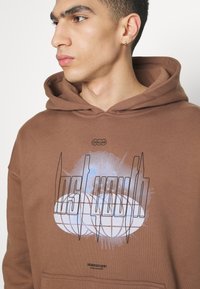 Felpa con cappuccio marrone in cotone con stampa grafica e accenti blu. Include un cappuccio con cordino e una tasca a canguro frontale.