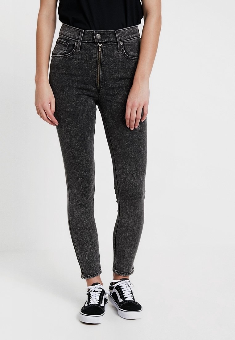 Jeans ajustados de tiro alto en mezclilla negra con cierre de cremallera y bolsillos delanteros, con textura descolorida y costuras en contraste. Combinados con zapatillas negras.