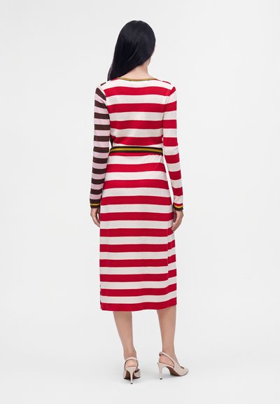 Femme debout, de dos, portant une robe midi à manches longues avec des rayures horizontales rouges et blanches et une manche rayée marron, talons beiges.