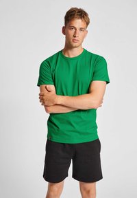 Grön bomull t-shirt med korta ärmar och rundad halsringning, kombinerad med svarta shorts. Tyget verkar slätt och designen är enkel och åtsittande.
