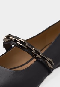 Μαύρο δερμάτινο επίπεδο παπούτσι με διακοσμητικό λαμπερό μαύρο και χρυσό λουράκι με αλυσίδα στην πάνω πλευρά.