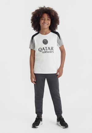 Naeratav laps lokkis juustega seisab, kandes valget ja halli Paris Saint-Germaini spordisärki, tumedaid pükse ning musti tenniseid ühevärvilise tausta ees.