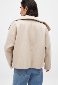 Beige wollen blend jas met een brede kraag en verlaagde schouders, met een boxy silhouet. Achteraanzicht toont schone lijnen en minimale details.