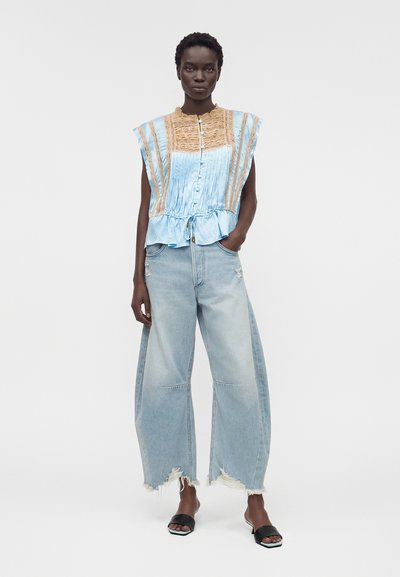 Γυναίκα φορούν μπλε και μπεζ αμάνικη μπλούζα με πιέτες και ανοιχτόχρωμα φθαρμένα φαρδιά τζιν, στέκεται με το ένα χέρι στην τσέπη και μαύρα σανδάλια.