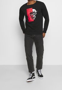Felpa nera con un design rettangolare rosso che presenta un grafico di un teschio. Abbinata a jeans grigi e scarpe sportive nere alte.