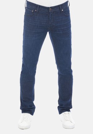 Jeans de mezclilla azul oscuro con un corte ajustado, que presentan un diseño tradicional de cinco bolsillos y un desvanecido sutil. Combinados con zapatillas blancas.