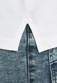 Weißes Baumwollhemd mit einem geteilten Saum hinten, kombiniert mit verblassten blauen Jeans aus Denim mit strukturiertem Material und kontrastierenden Nähten.