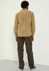 Kariertes Khaki-Shirt aus Cord mit einem Rückenfalte, langen Ärmeln und Knopfmanschetten; kombiniert mit dunkelbraunen Cargo-Hosen mit Seitentaschen.