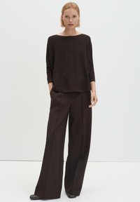 Frau mit kurzen blonden Haaren, die einen dunkelbraunen, locker sitzenden Pullover und weit geschnittene Hosen trägt, vor einem schlichten hellen Hintergrund stehend.