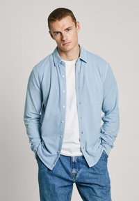 Camisa de manga larga de color azul claro, hecha de una tela suave, con botones en la parte frontal, diseño con cuello, y combinada con una camiseta blanca y jeans azules.