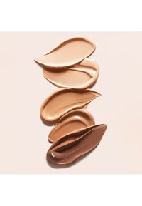 Cinque campioni di fondotinta in diverse tonalità di beige e marrone su uno sfondo chiaro.