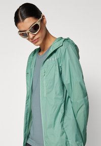 Lightweight mint green rain jacket with a hood, zippered chest pocket, and breathable fabric, layered over a light blue t-shirt.
