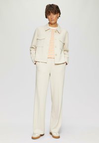 Blazer texturé ivoire avec deux poches poitrine, assorti à un pantalon à jambes larges. Chemise rayée orange et blanche en dessous, coupe décontractée.