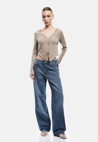 Beige Rippstrick-Cardigan mit Spitzenbesatz, kombiniert mit blauen, weiten Jeans. Das Outfit hat ein kurzes Design und eine lässige Passform aus Denim.