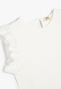 Top blanco de manga corta que presenta un adornado dobladillo con volantes de ojales en un hombro y un escote redondo con cierre en forma de ojal en la parte posterior.
