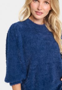 Mörkblått fluffigt tröja med rund halsringning och korta ärmar. Texturen är mjuk och plyschig, vilket ger djup åt designen.