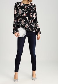 Blouse noire à fleurs avec manches longues, ornée de fleurs blanches et roses. Associée à un jean skinny foncé et des chaussures à talons argentées.
