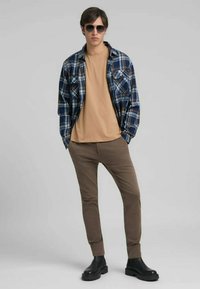 Joven con gafas de sol, camisa a cuadros azul sobre una camiseta beige, pantalones marrones y botas negras, de pie contra un fondo gris liso.