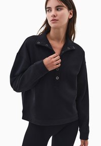 Sudadera negra con un cuello alto, tapeta abotonada y herrajes plateados. Corte holgado con mangas largas y puños acanalados, confeccionada en una tela suave.