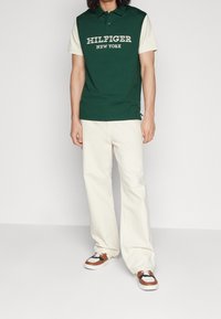 Mann in grün-creamfarbenem kurzärmligen Poloshirt mit dem Schriftzug "Hilfiger New York", creamfarbenen weitgeschnittenen Hosen und braun-weißen Sneakers steht.