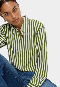 Camicia a righe verde e bianca con colletto classico e maniche lunghe, realizzata in un tessuto liscio. Abbinata a jeans in denim blu.