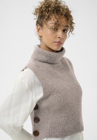 Barna színű kötött garbó mellény bordázott textúrával és három sötétbarna gombbal a bal oldalon, fehér ing fölé rétegezve.