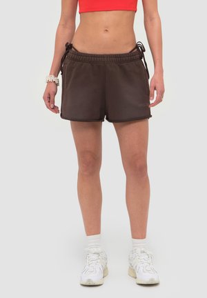 Personne portant un short à cordon de serrage brun foncé, des baskets blanches, des chaussettes blanches et un top court rouge sur un fond clair uni.