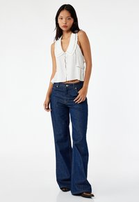 Vit ärmlös blus med en stor krage och knäppning fram, ihop med mörkblå utsvängda jeans. Tyget verkar slätt och strukturerat.