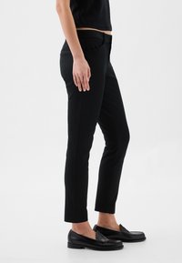 Femme portant un pantalon noir slim-fit et des mocassins noirs, debout de profil sur un fond clair uni.