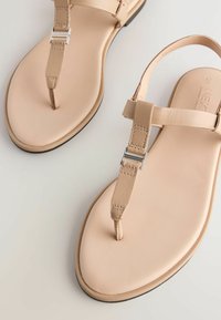 Beige leather sandals featuring a thong design, flat sole, and metallic accents on straps. Smooth texture with minimal stitching details.