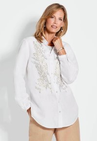 Camicia bianca in lino con colletto, chiusura a bottoni sul davanti e decorazioni dorate sul lato sinistro. Maniche lunghe con bottoni sui polsini.