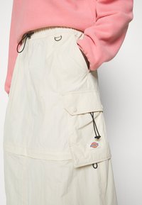 Falda de color beige claro, hecha de tejido texturizado, que cuenta con una cinturilla elástica, bolsillos laterales tipo cargo y un parche con un logo circular.