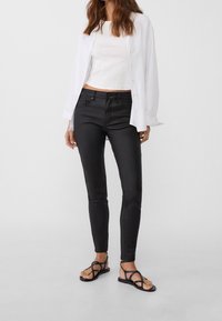 Černé skinny džíny s hladkou texturou, doplněné o přiléhavý bílý crop top a volnou bílou košili na knoflíky, spárované s černými sandály.