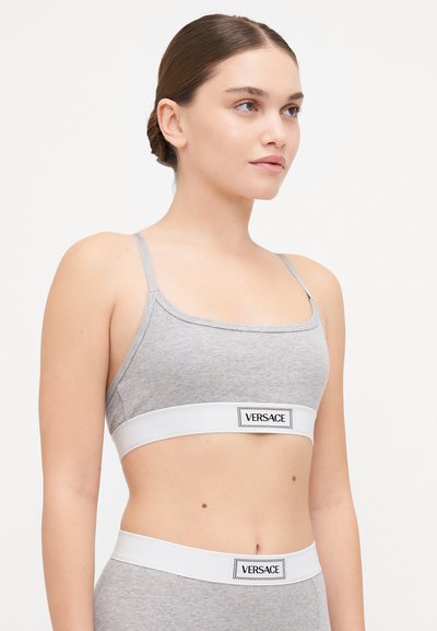 Pelēkā ribotā auduma bralette ar plānām regulējamām siksnām un baltu elastīgu joslu, uz kuras ir "VERSACE" logo. Vienkāršs un minimālistisks dizains.