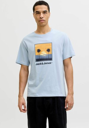 Homme portant un T-shirt bleu clair avec un coucher de soleil, deux palmiers et le texte "Jack & Jones", accompagné d'un pantalon noir, debout devant un fond uni.