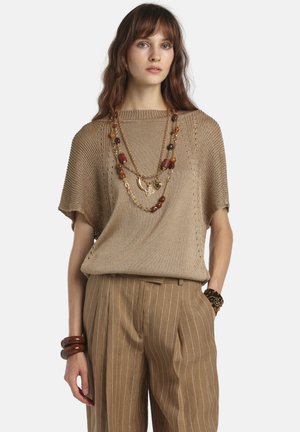 Kvinde iført en løs, strikket beige top, bukser med brede ben og nålestriber, lag-på-lag perle- og kædehalskæder samt chunky armbånd på begge håndled.