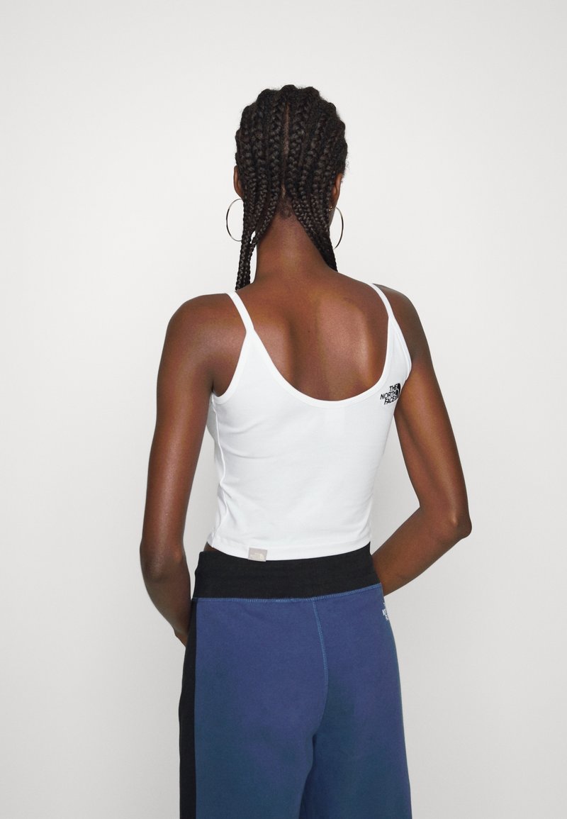 Femme avec des cheveux tressés portant un haut blanc sans manches et un pantalon bleu, debout les mains dans le dos devant un fond uni.