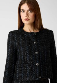 Giacca di tweed texturizzata nera e blu con scollo rotondo, cinque bottoni d'argento e due tasche finte.