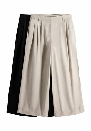 Sötét hajú nő fekete cipzáras pulóverben, krémszínű, bő szárú nadrágban, fekete övvel és fekete belebújós cipővel, karját keresztbe téve áll egy fehér fal előtt.