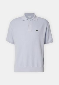 Ljusblå polo skjorta i mjukt tyg, med ribbad krage, korta ärmar, två knappar och en grön Lacoste-logotyp på bröstet.