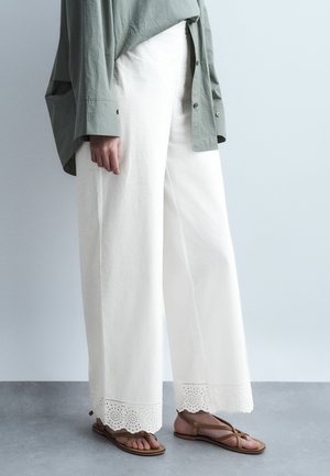 Person iført hvide bukser med vide ben og øjelynlacekant, olivengrøn skjorte med knapper og brune flade sandaler med stropper, stående på gråt gulv.